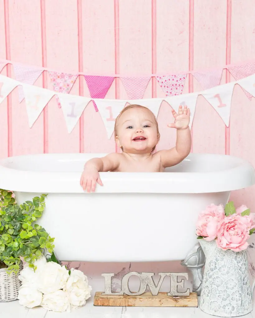 a little girl waves from a bath tub at her first birthday tub splash photo shoot