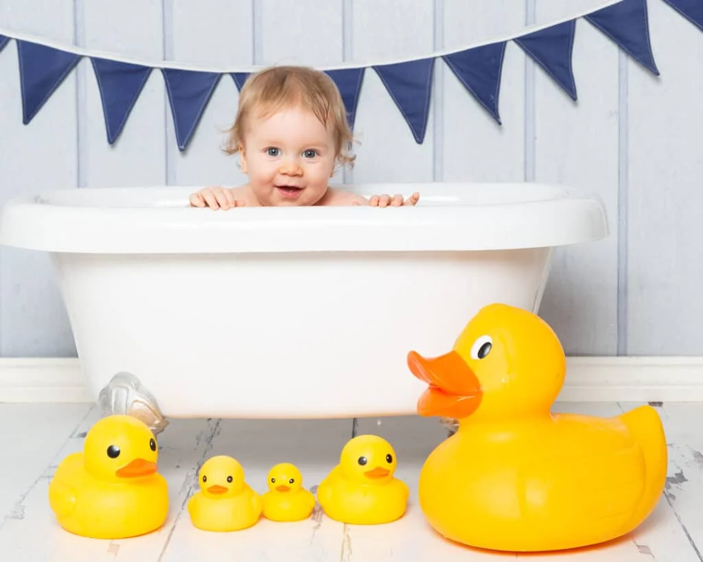 Baby Photography in East Grinstead West Sussex a little boy poses in a bath tub holding onto the side with some brightly coloured rubber ducks at a first birthday photo shoot