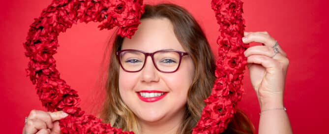 a woman poses against a red background looking through a flower heart shape
