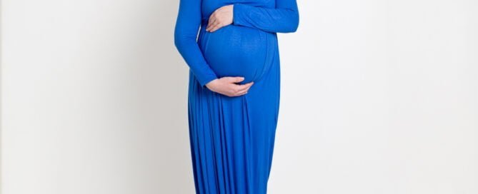 A full length picture of a pregnant woman posed in a photography studio against a white backdrop. She is looking down at her bump and wearing a long blue maternity dress.