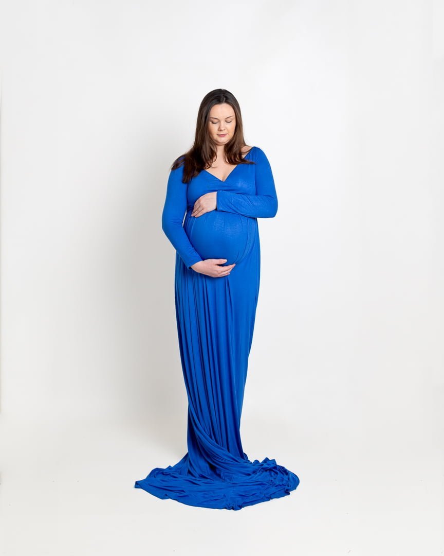 A full length picture of a pregnant woman posed in a photography studio against a white backdrop. She is looking down at her bump and wearing a long blue maternity dress.