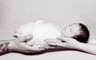 A black and white image of a swaddled sleepy newborn being held in both parents hands