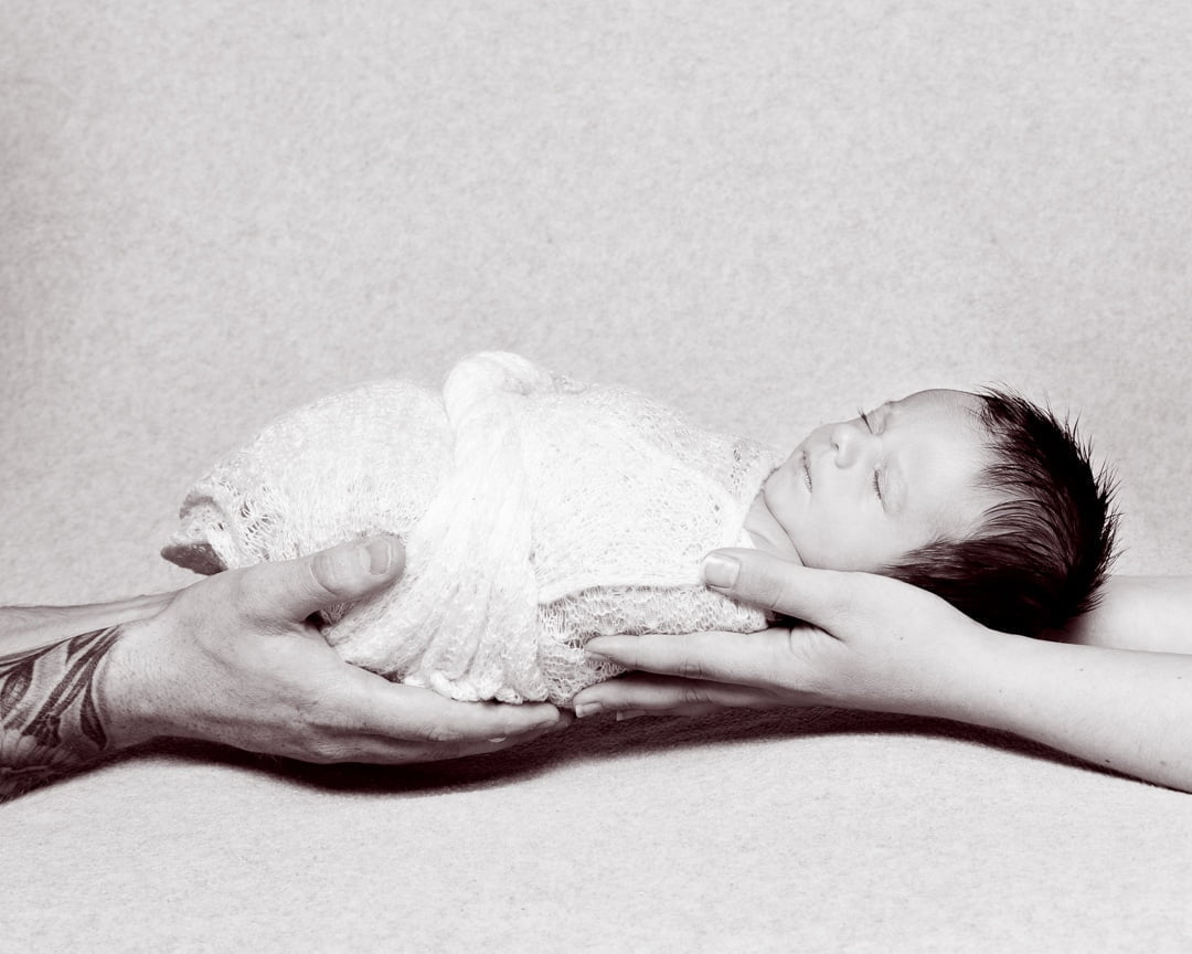 A black and white image of a swaddled sleepy newborn being held in both parents hands