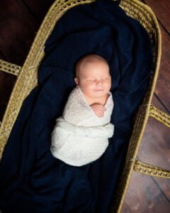 A newborn baby is swaddled in a white wrap while sleeping in a Moses Basket padded with a navy blue blanket