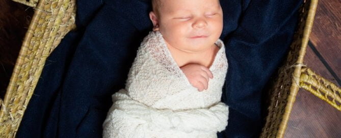 A newborn baby is swaddled in a white wrap while sleeping in a Moses Basket padded with a navy blue blanket