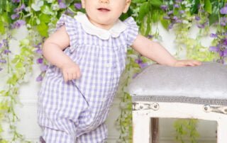 a little girl in a purple and white romper poses in front of a flower wall at a photo shoot