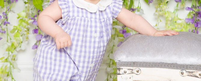 a little girl in a purple and white romper poses in front of a flower wall at a photo shoot