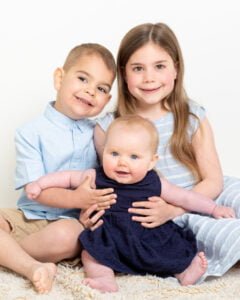 An older brother and sister support their younger sibling against a white background at a professional family portrait shoot