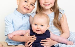 Two older siblings support their baby sister with all three dressed in shades of blue