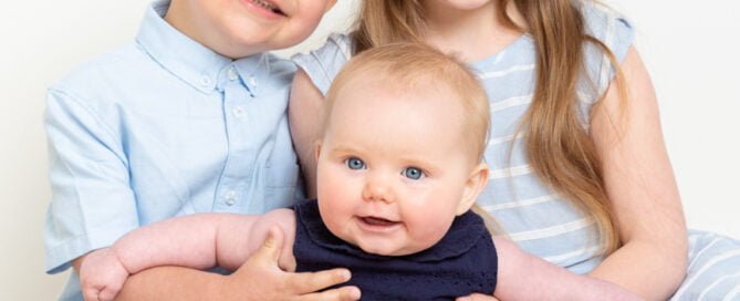 Two older siblings support their baby sister with all three dressed in shades of blue