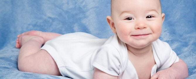 a baby in a white vest smiles at the camera while lying on his tummy on a blue blanket