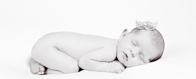 a black and white newborn image of a sleeping baby tucked up on their tummy and wearing a tiara
