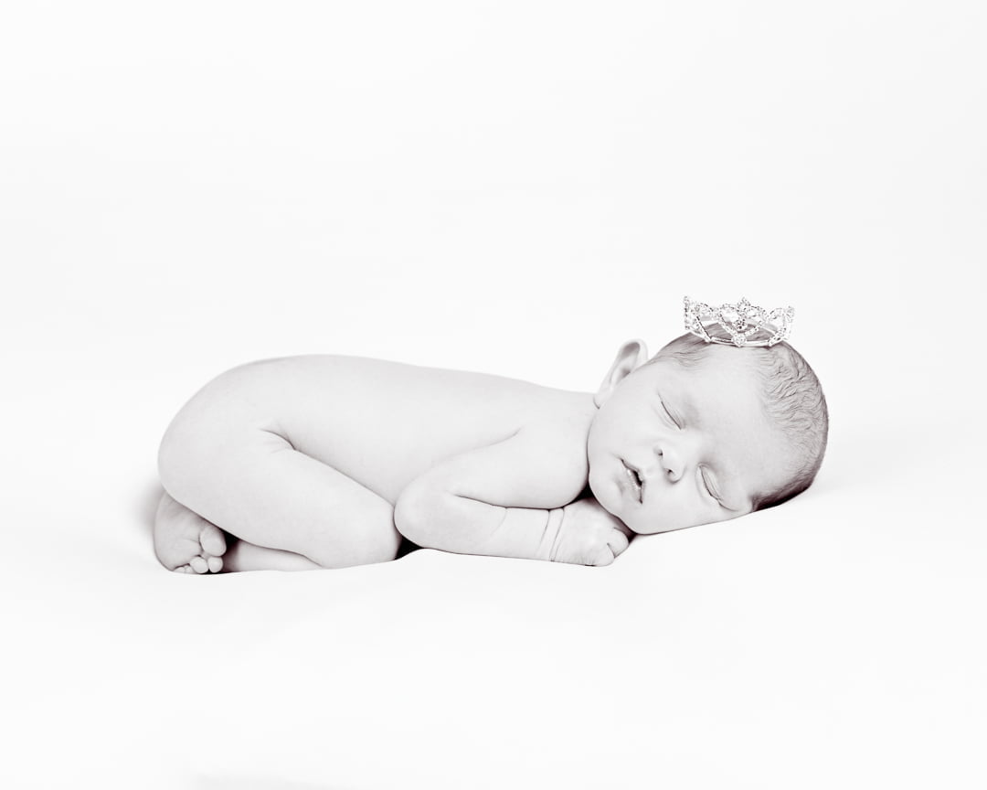 a black and white newborn image of a sleeping baby tucked up on their tummy and wearing a tiara