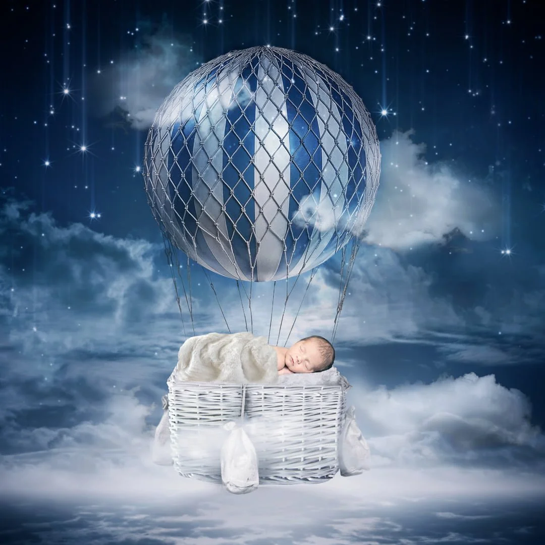 A digitally composite image of a sleeping newborn tucked up under a blanket on a hot air balloon in the night sky