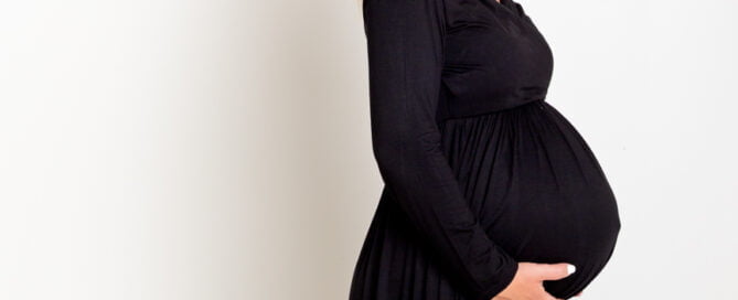 A pregnant woman smiles for the camera against a neutral background whilst wearing a long flowing black dress