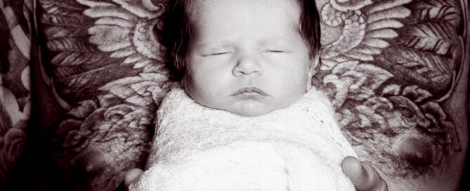 a black and white image of a dad holding his newborn to his chest while his tattoo frames the baby with angel wings