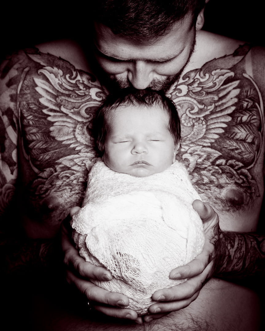 a black and white image of a dad holding his newborn to his chest while his tattoo frames the baby with angel wings