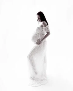 A pregnant woman is posing against a white background in a flowing white maternity dress. A black and white image taken at my studio in East Grinstead.