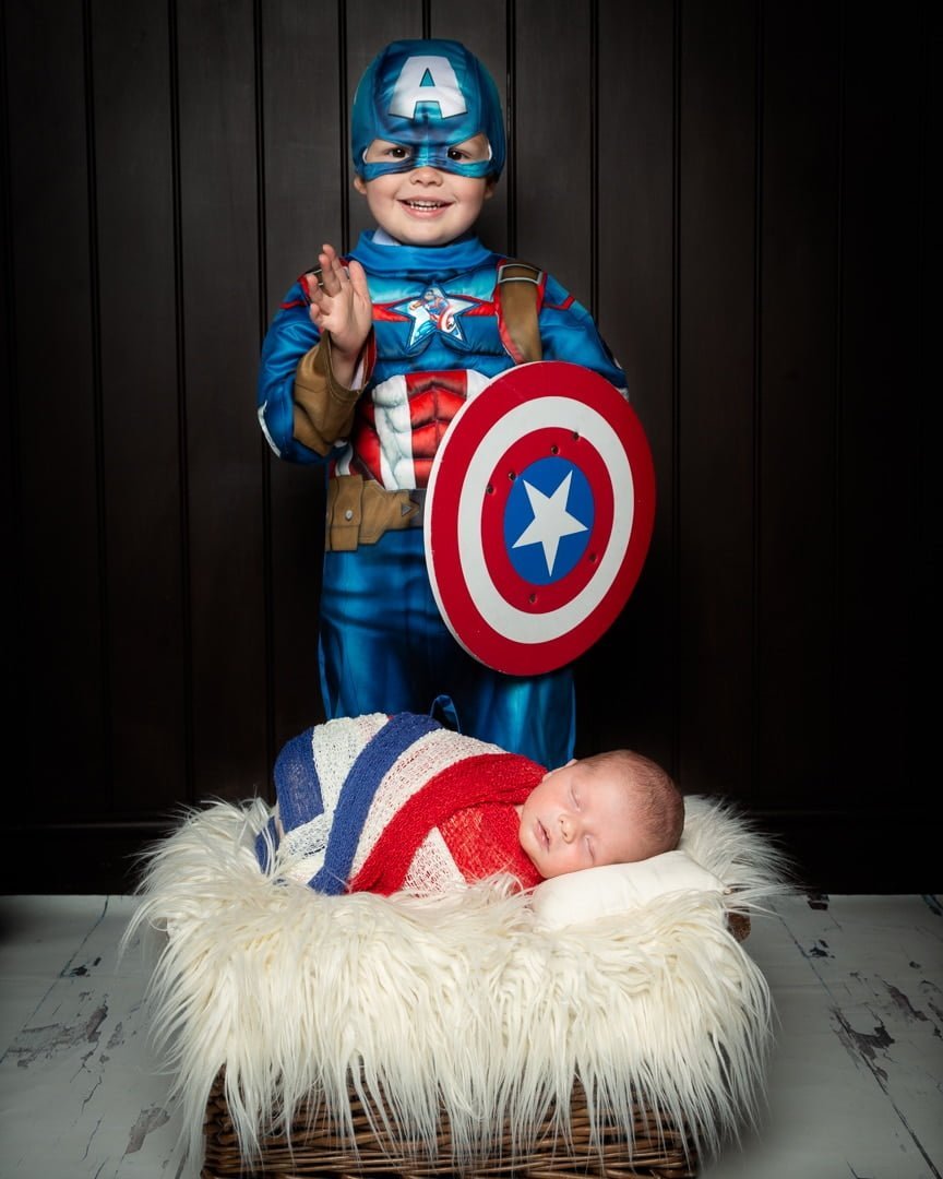 a little boy dressed as Captain America poses with his newborn brother wrapped in red, white and blue