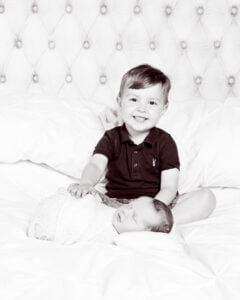 A newborn baby and their big brother pose on a bed together for a professional photo shoot - a black and white image taken in my East Grinstead Studio