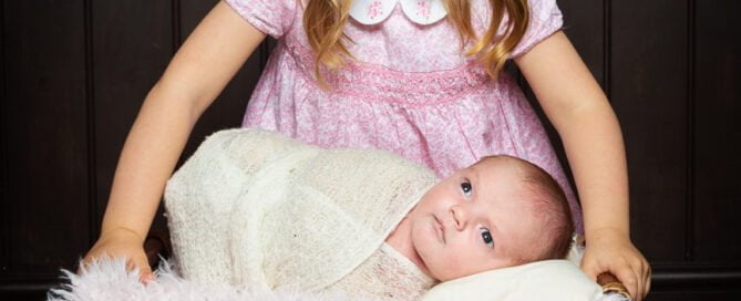a big sister looks down at their newborn sibling swaddled in white