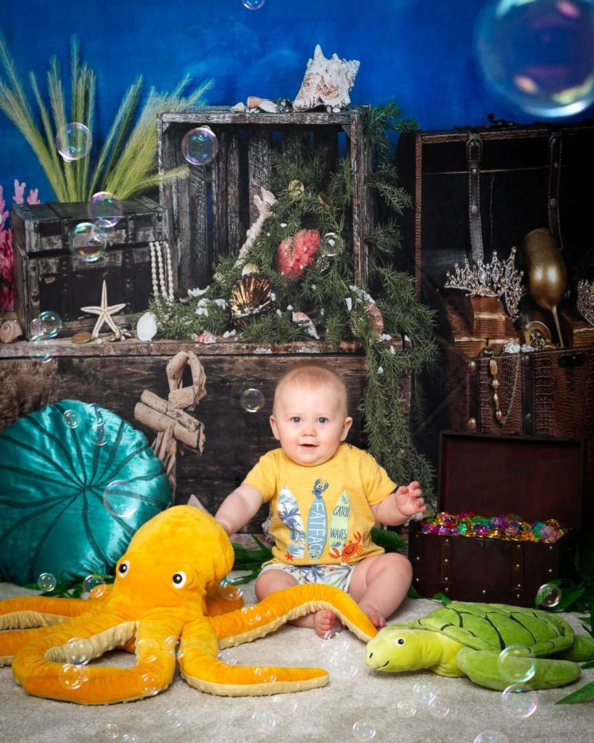a baby posing in an underwater scene set up in a photographic studio with a backdrop and plush props