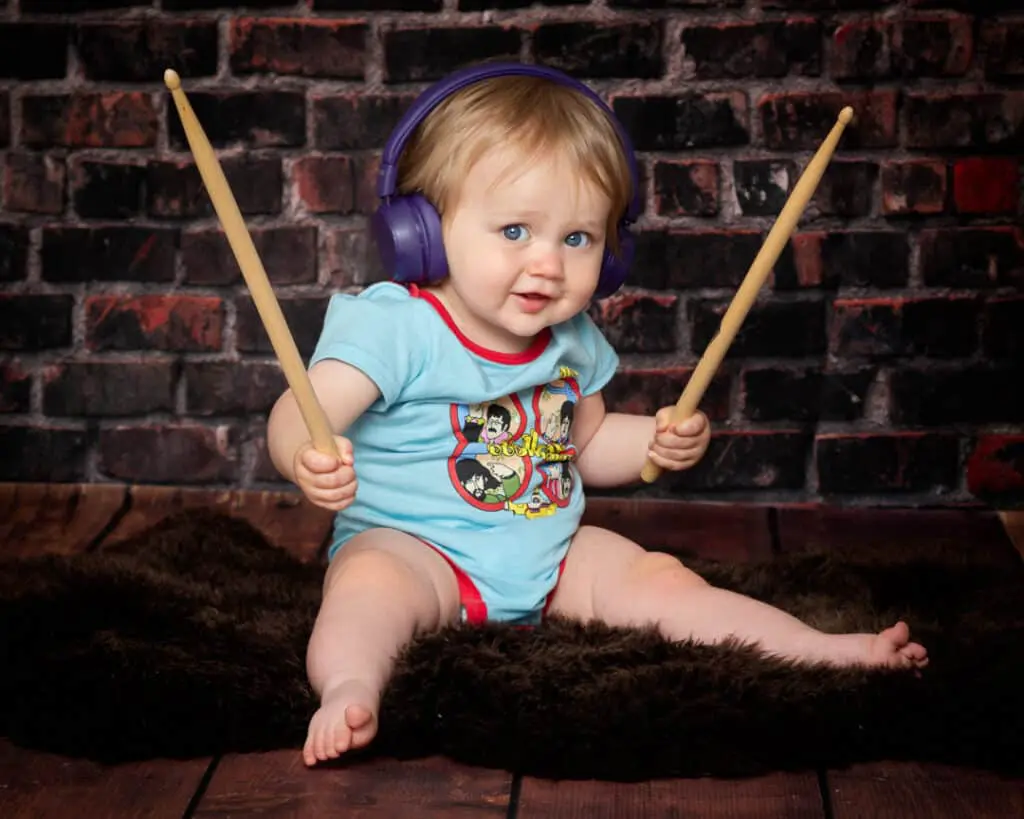 a baby sits in front of a brick wall in a beatles vest holding drumsticks and wearing headphones