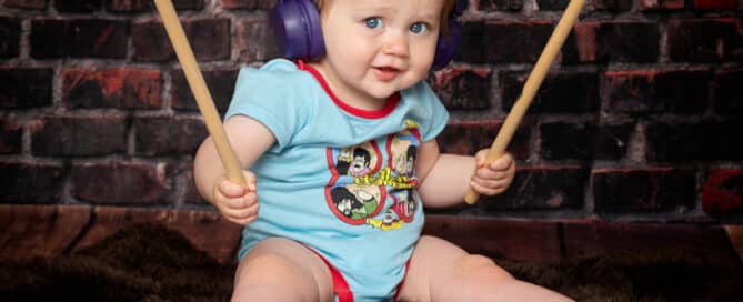 a baby sits in front of a brick wall in a beatles vest holding drumsticks and wearing headphones