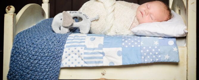 A newborn sleeping on my miniature white bed prop with a blue quilt and blanket