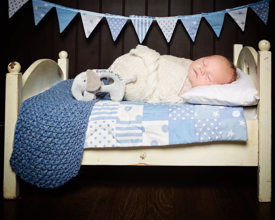 A newborn sleeping on my miniature white bed prop with a blue quilt and blanket