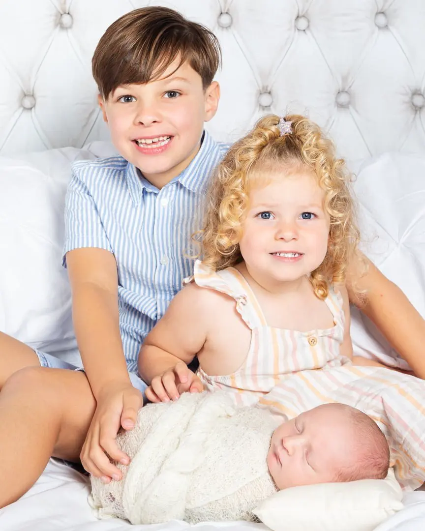 Newborn Photography East Grinstead West Sussex Three children sitting on a bed looking at the camera, an older brother and sister and a swaddled newborn