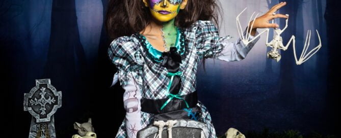 a little girl in spooky face paint poses at a halloween themed photo shoot in a graveyard set