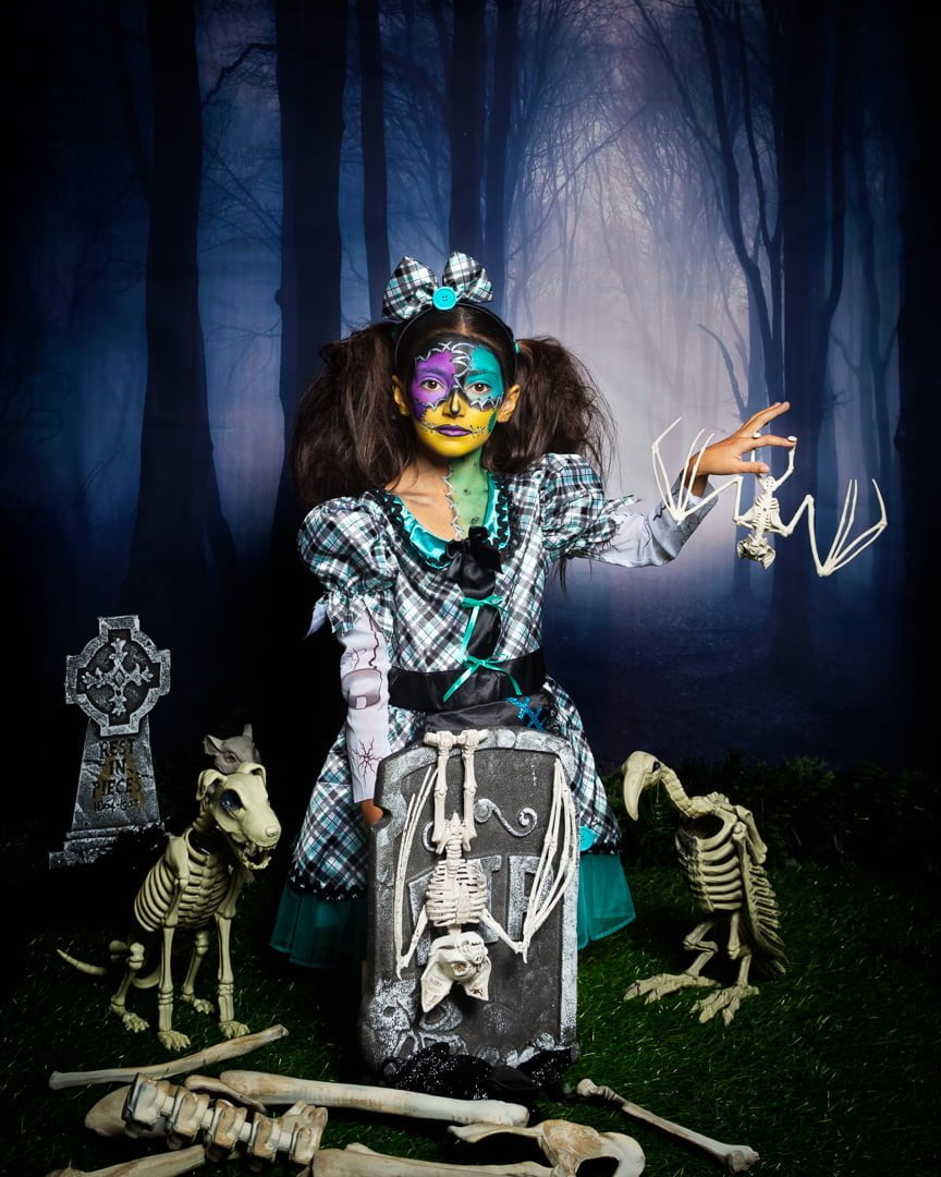 a little girl in spooky face paint poses at a halloween themed photo shoot in a graveyard set
