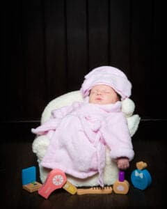 A newborn baby wearing a pink robe and head towel relaxes in a prop spa chair surrounded by beauty products