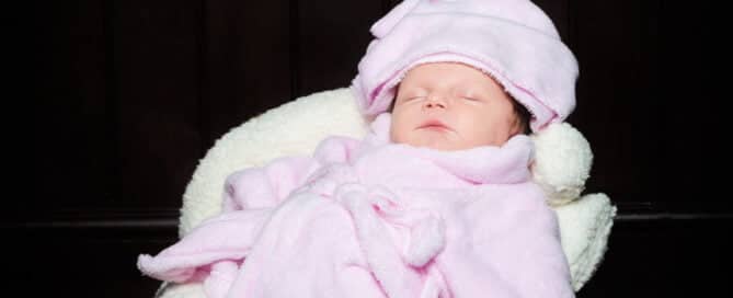 A newborn baby wearing a pink robe and head towel relaxes in a prop spa chair surrounded by beauty products