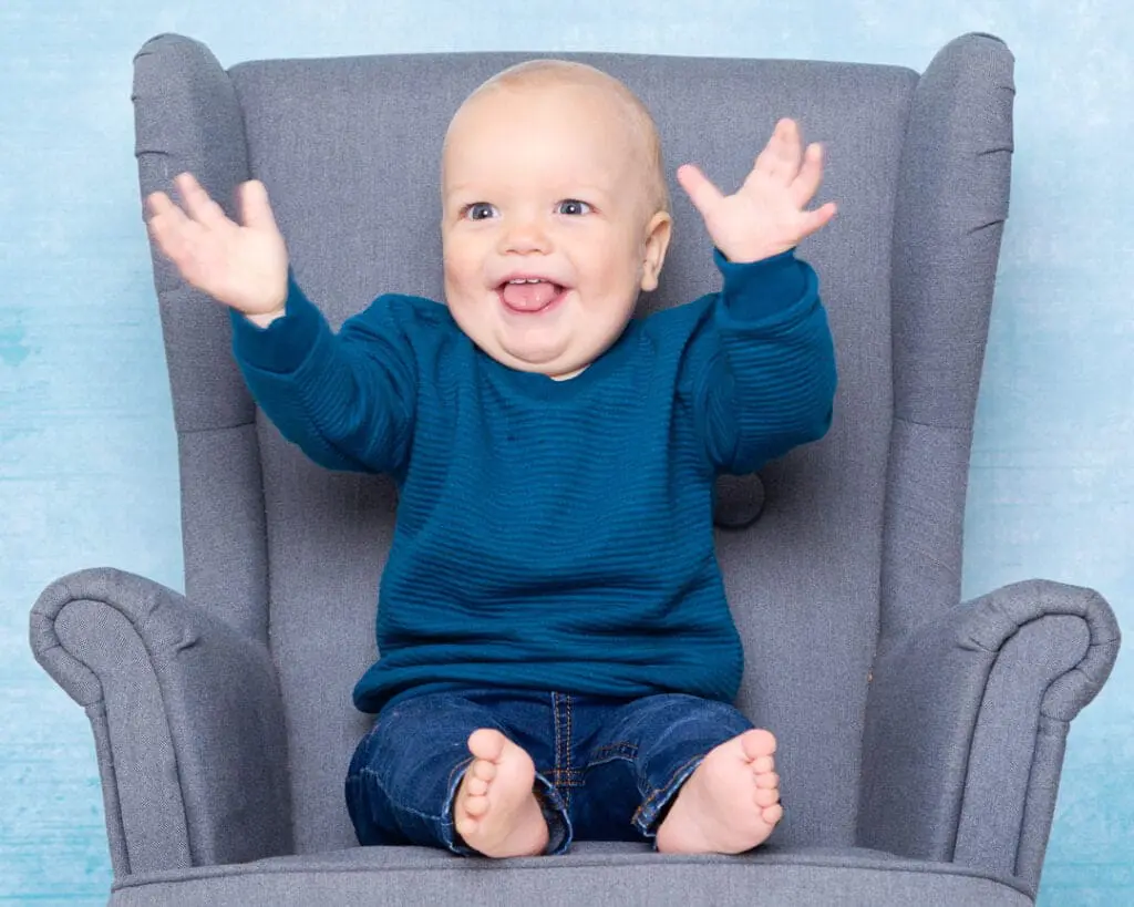 a little boy in a blue jumper smiles and claps his hands for a photo in a studio
