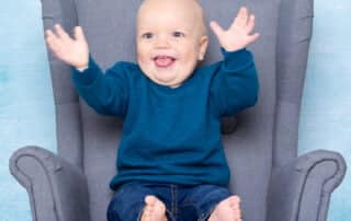 a little boy in a blue jumper smiles and claps his hands for a photo in a studio
