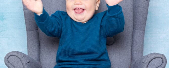 a little boy in a blue jumper smiles and claps his hands for a photo in a studio