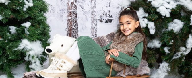 a little girl relaxes on a wooden sledge in a snow themed christmas photo shoot set