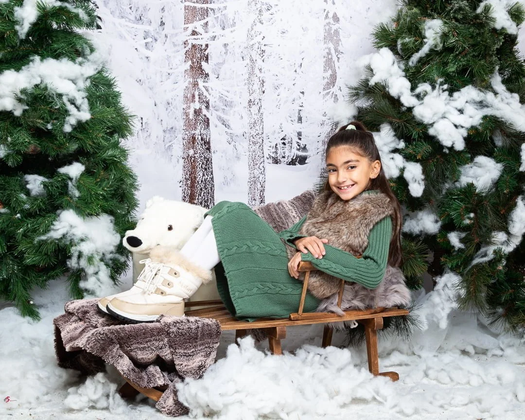 a little girl relaxes on a wooden sledge in a snow themed christmas photo shoot set