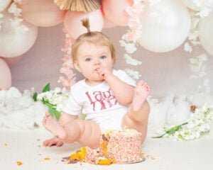  A little girl sits behind a cake covered in sprinkles tucking into some icing at her cake smash first birthday photo shoot