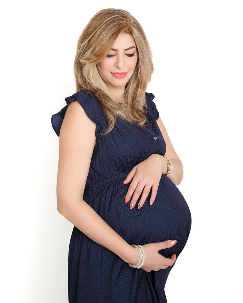 A woman poses in the studio in a blue tight fitting dress against a white background in my photographic studio She is pregnant and has her arms wrapped around her bump