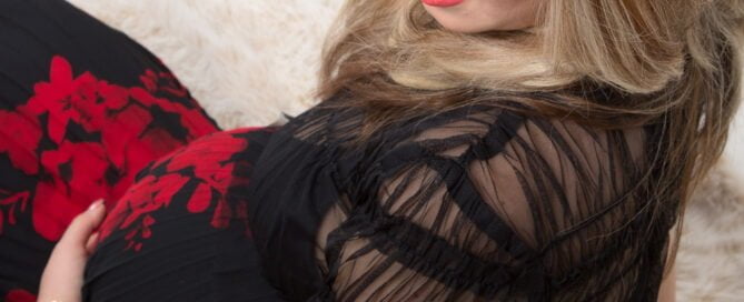 A pregnant woman wearing a red and black outfit holds her bump as she sits on the floor and looks down over her shoulder