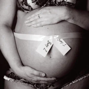 A pregnant lady cradles her bare bump in a black and white image. The bump is wrapped in a white ribbon with two labels reading 'baby 1' and 'baby 2'