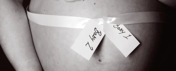 A pregnant lady cradles her bare bump in a black and white image. The bump is wrapped in a white ribbon with two labels reading 'baby 1' and 'baby 2'