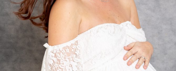 a pregnant woman poses against a grey canvas background looking down at her bump and wearing a white lace maternity dress