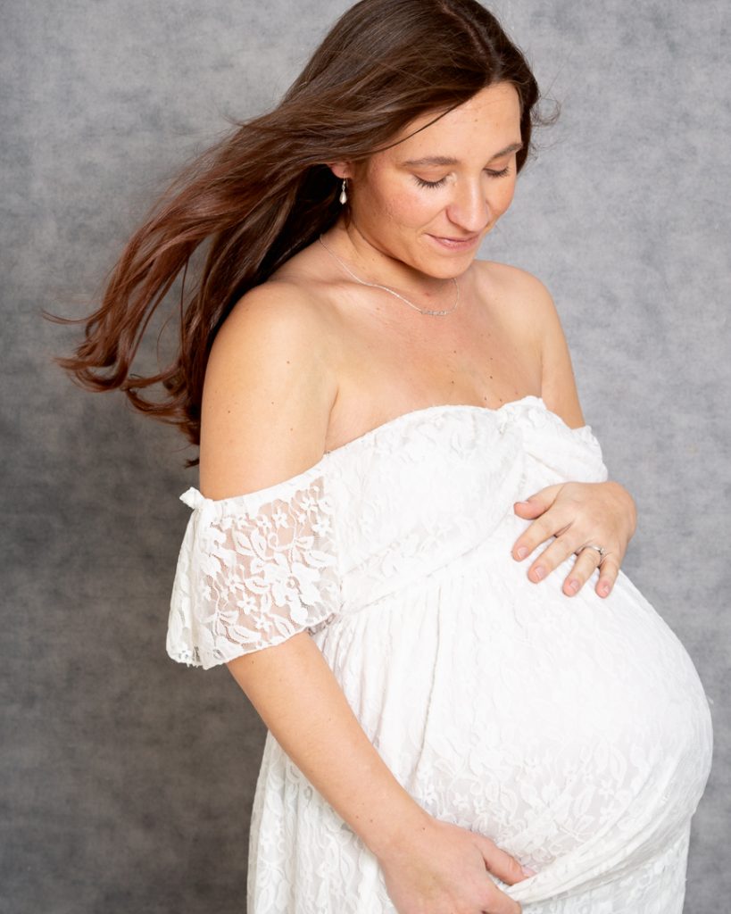 a pregnant woman poses against a grey canvas background looking down at her bump and wearing a white lace maternity dress