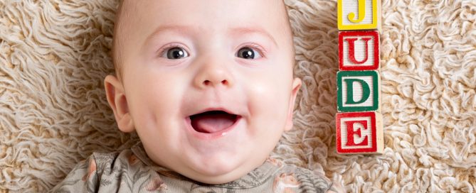 A smiling baby lying on his back looking up at the camera next to his name spelt out in alphabet letter blocks