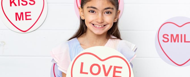 a little girl poses with a giant love heart themed candy sign that says I love you at a Mother's Day Photo Shoot