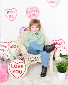 a little boy sits on a white chair surrounded by Mother's Day props